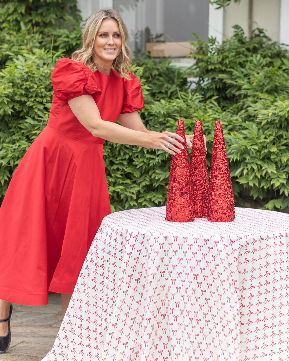 Red bow print round tablecloth styled with festive red dress and sequined decorations, outdoor setting.