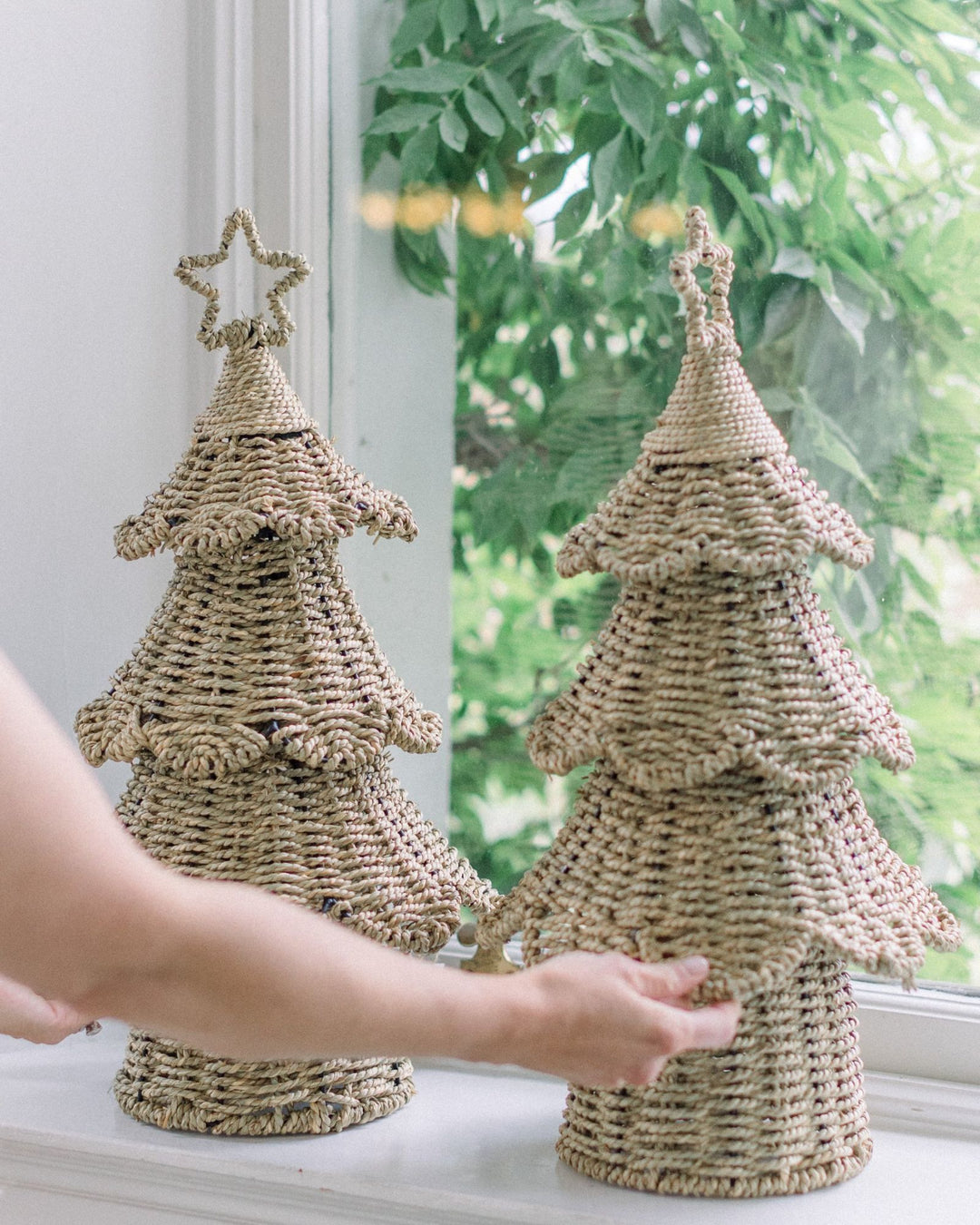 Two handwoven rattan Christmas trees with stars styled on a windowsill with greenery.