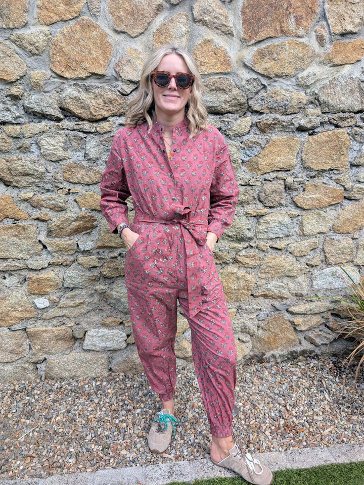 Model wearing maroon handcrafted long-sleeve cotton jumpsuit standing outdoors against a stone wall.