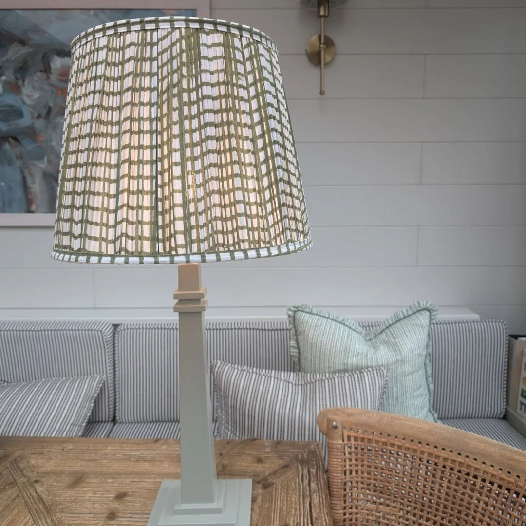 Green grid pleated empire lampshade styled in a cosy living room setting with striped sofa and soft lighting.