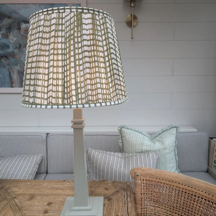Green grid pleated empire lampshade styled in a cosy living room setting with striped sofa and soft lighting.