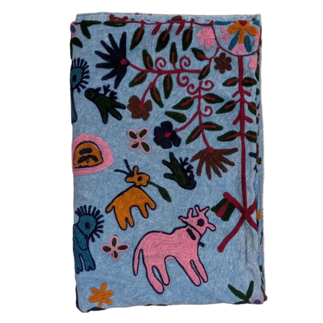 A folded light blue blanket or throw featuring a colorful hand-stitched folk art pattern with pink and orange animals, black and red floral motifs, and green leaves on a white background.